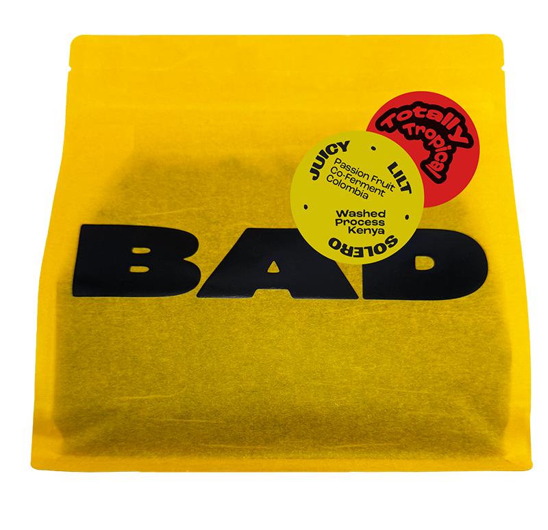 A bright yellow stand-up pouch of Passion fruit co-fermented coffee with large black 'BAD' lettering and circular coffee origin stickers reading Juicy, Lilt, Solero and "Washed Process Kenya x Passion Fruit Co-Ferment Colombia".
