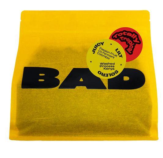 A bright yellow stand-up pouch of Passion fruit co-fermented coffee with large black 'BAD' lettering and circular coffee origin stickers reading Juicy, Lilt, Solero and "Washed Process Kenya x Passion Fruit Co-Ferment Colombia".