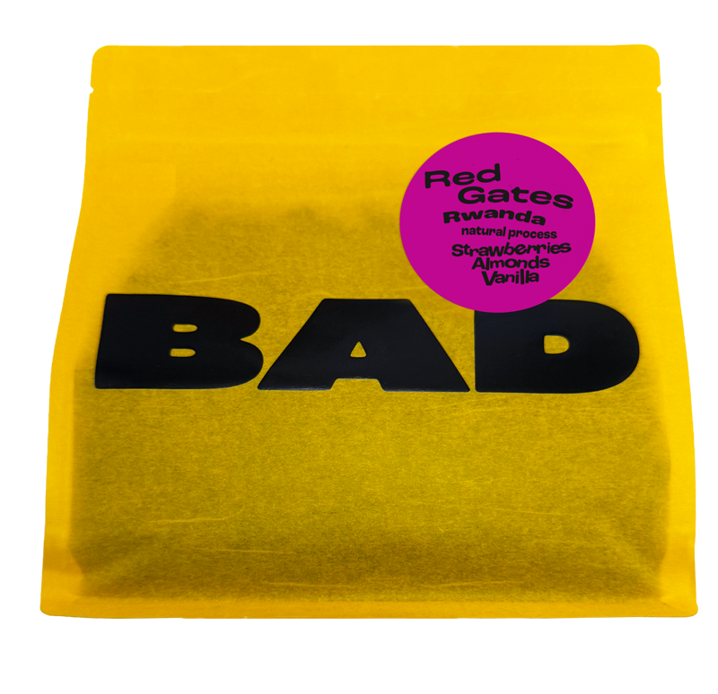 A bright yellow bag of coffee with large black 'BAD' lettering and a circular magenta label reading 'Red Gates Rwanda natural process Strawberries Almonds Vanilla'.