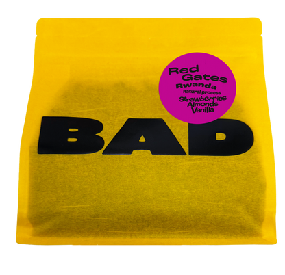 A bright yellow bag of coffee with large black 'BAD' lettering and a circular magenta label reading 'Red Gates Rwanda natural process Strawberries Almonds Vanilla'.