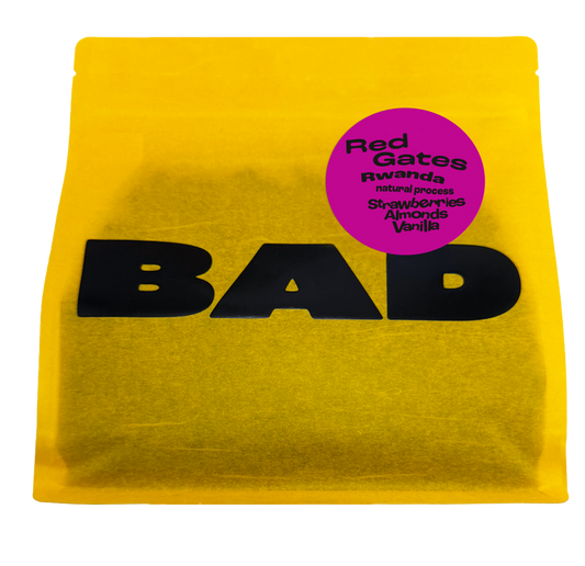 A bright yellow bag of coffee with large black 'BAD' lettering and a circular magenta label reading 'Red Gates Rwanda natural process Strawberries Almonds Vanilla'.