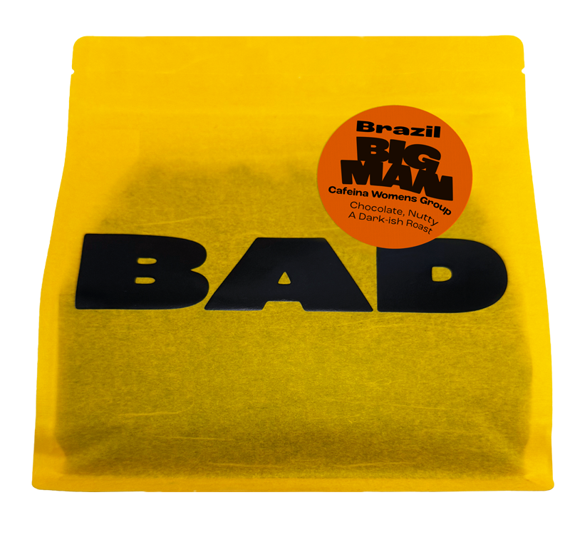 A bright yellow bag of coffee with bold black 'BAD' lettering and a circular orange sticker reading 'Brazil BIG MAN Cafeína Womens Group Chocolate, Nutty, A Dark-Rich Roast'.