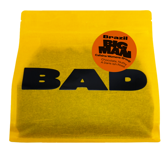 A bright yellow bag of coffee with bold black 'BAD' lettering and a circular orange sticker reading 'Brazil BIG MAN Cafeína Womens Group Chocolate, Nutty, A Dark-Rich Roast'.