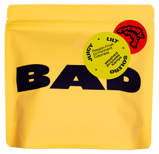 A yellow stand-up pouch of coffee with large black 'BAD' lettering and two circular flavor stickers reading 'Totally Tropicale' and 'Passion Fruit Co-Fermented coffee'.
